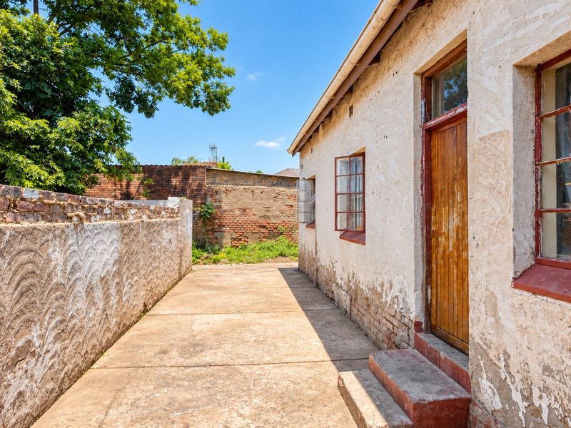 Rear exterior view showing a concrete pathway, weathered brick boundary wall, and the side facade of the house with original steel-framed windows.