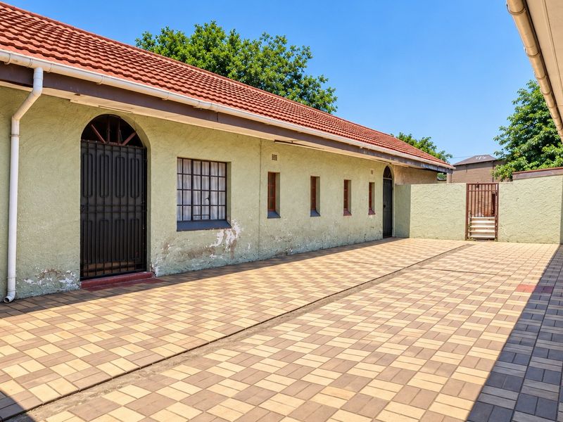 A paved rear courtyard area featuring a long building facade with terracotta roof tiles, security gates, and security bars on the windows.