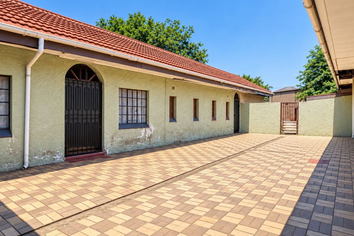 A paved rear courtyard area featuring a long building facade with terracotta roof tiles, security gates, and security bars on the windows.