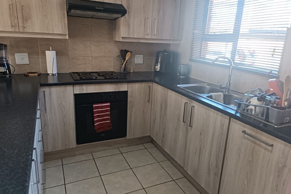 A functional kitchen featuring light wood-grain cabinetry, black granite countertops, built-in oven, electric hob, and a stainless steel double sink with tiled flooring.