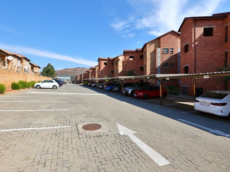 Exterior view of an apartment complex featuring a paved parking area with covered bays and brick building facades.
