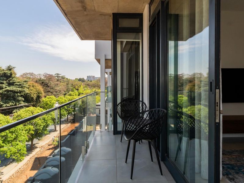 A modern balcony featuring a sleek glass balustrade, tiled flooring, and large sliding doors, offering a pleasant view of the surrounding greenery.
