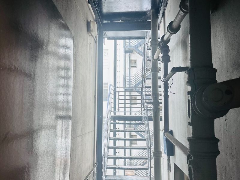 A narrow utility corridor featuring exposed plumbing and access to an external metal fire escape staircase.