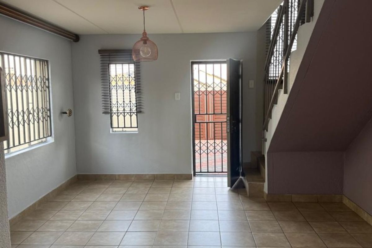 An open-plan living area featuring tiled flooring, a staircase, and security gates on windows and the entrance door.