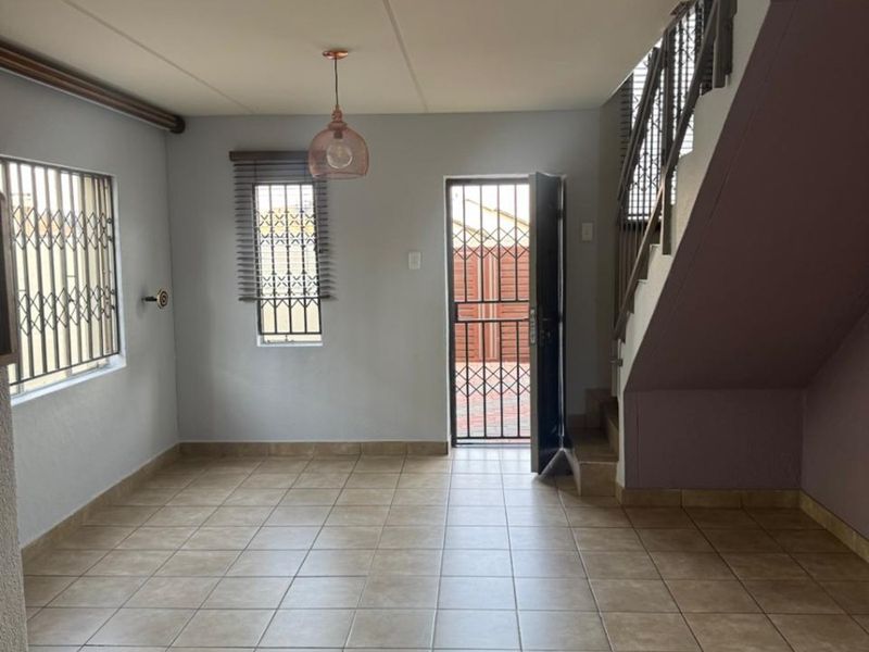 An open-plan living area featuring tiled flooring, a staircase, and security gates on windows and the entrance door.