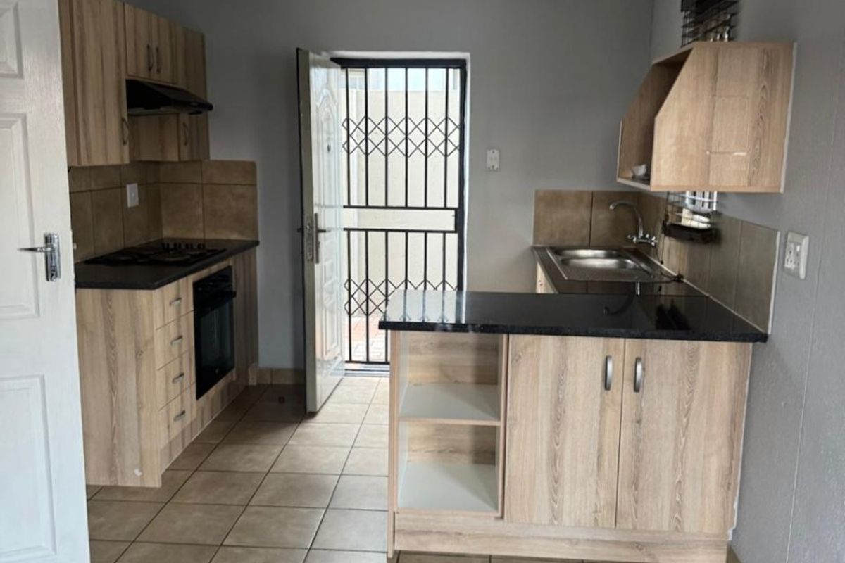 A functional kitchen featuring light wood-finish cabinetry, dark countertops, built-in oven and hob, and tiled flooring with direct access to an exterior security gate.