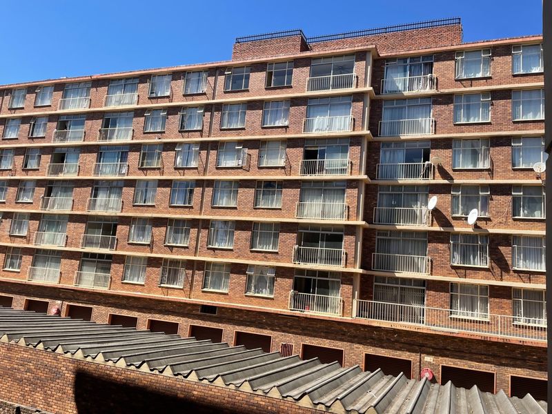 A multi-story apartment building exterior featuring a classic brick facade, individual balconies with metal railings, and a corrugated roof structure at the base.