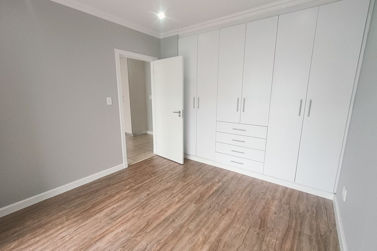 A bright, modern bedroom featuring neutral grey walls, high-quality laminate flooring, and extensive floor-to-ceiling built-in cupboards for ample storage.