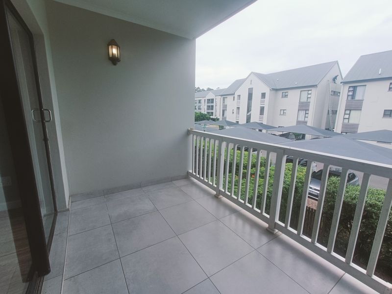 A clean, covered balcony featuring tiled flooring, a white railing, and a wall-mounted light fixture, overlooking the apartment complex parking area.