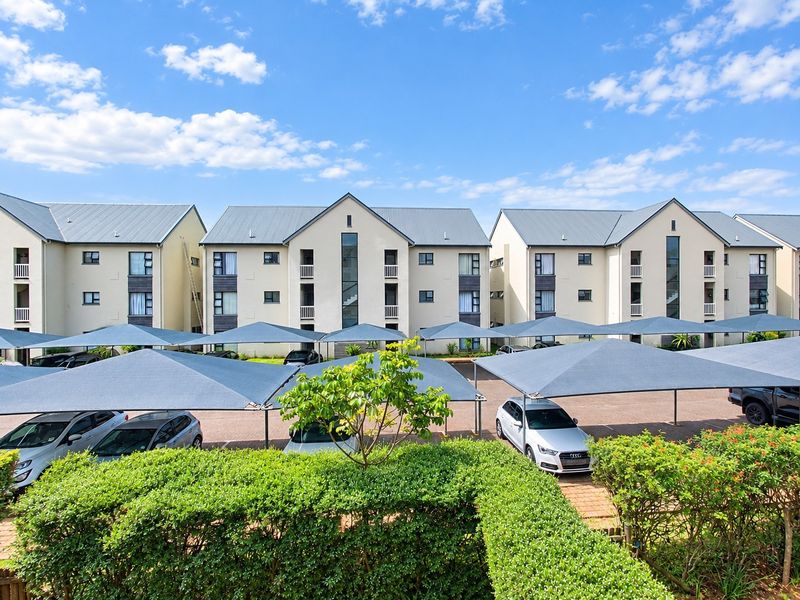 Exterior view of a modern apartment complex featuring multiple buildings with metal roofs, balconies, and covered parking bays set within a landscaped environment.