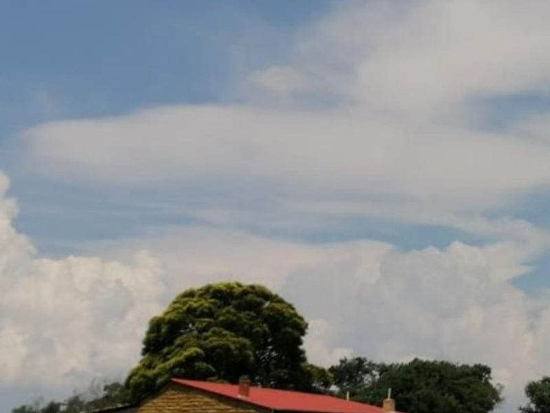 A rustic stone farmhouse featuring a red corrugated metal roof and an external water storage tank, set within a rural landscape.