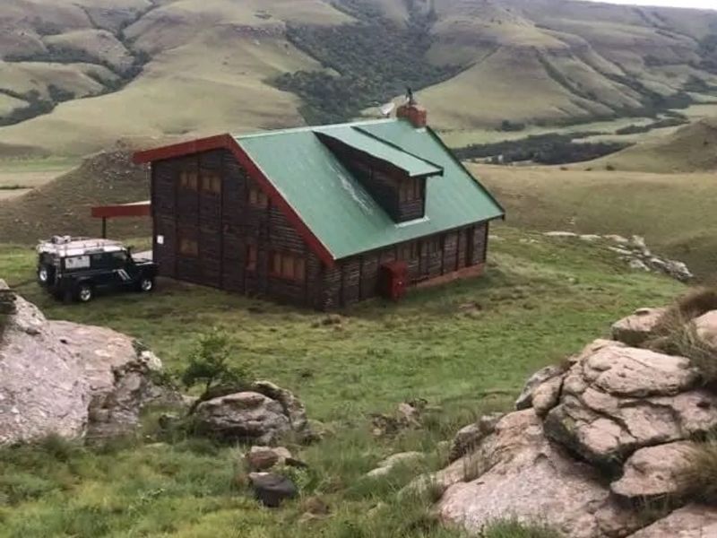 A rustic log cabin situated in a mountainous landscape, featuring a green metal roof and a prominent chimney.