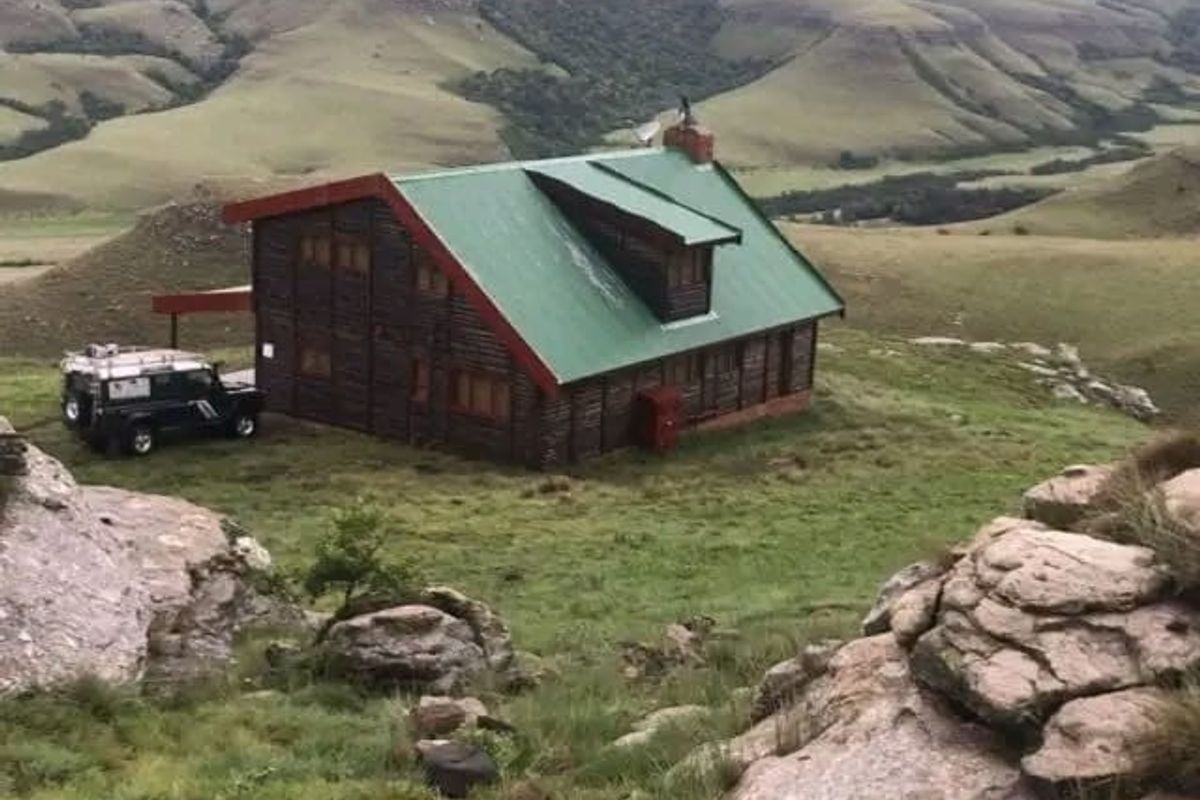 A rustic log cabin situated in a mountainous landscape, featuring a green metal roof and a prominent chimney.