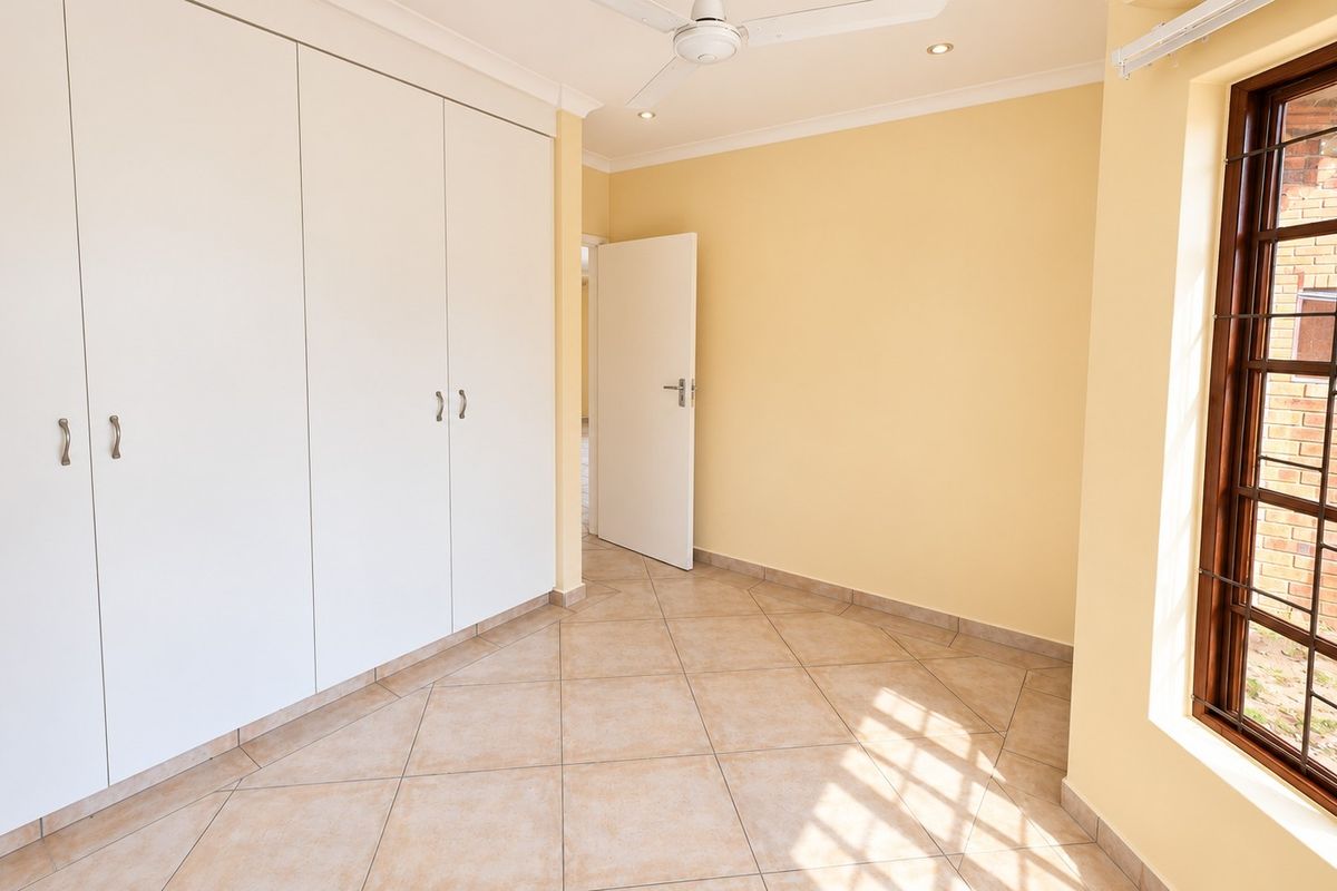 A bright, empty bedroom featuring tiled flooring, built-in cupboards, a ceiling fan, and a window with security bars.