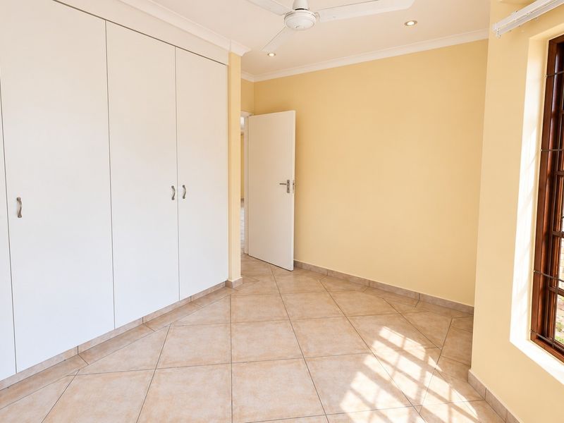 A bright, empty bedroom featuring tiled flooring, built-in cupboards, a ceiling fan, and a window with security bars.