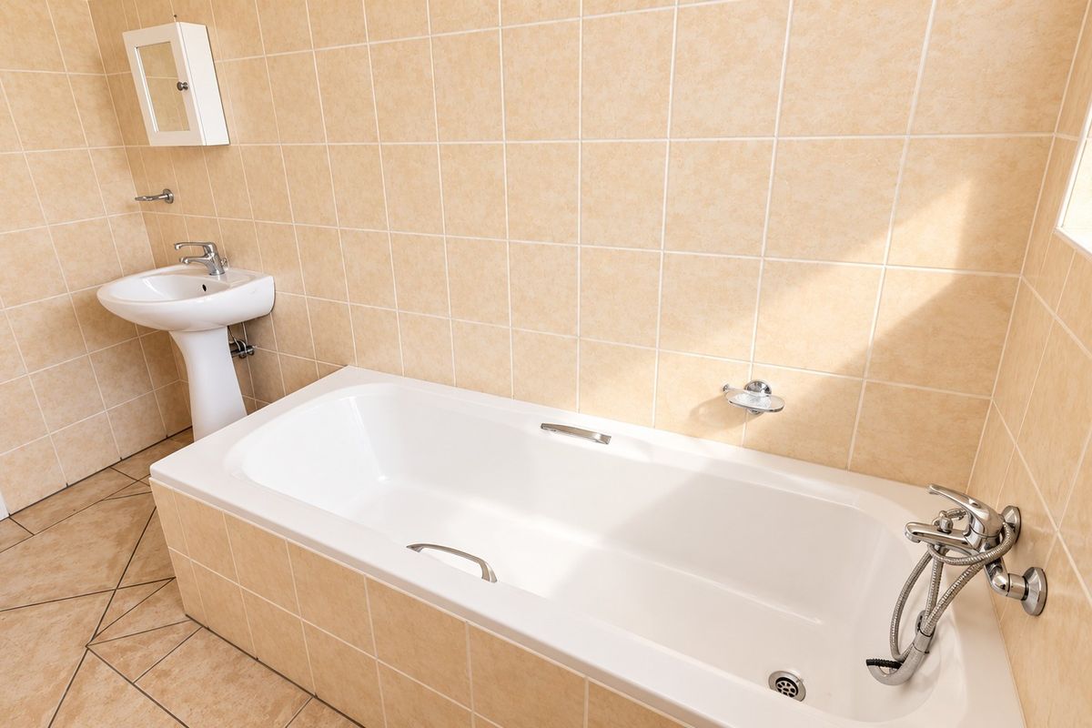 A clean, functional bathroom featuring a built-in bathtub with a handheld shower mixer, a pedestal basin, and neutral ceramic wall and floor tiling.