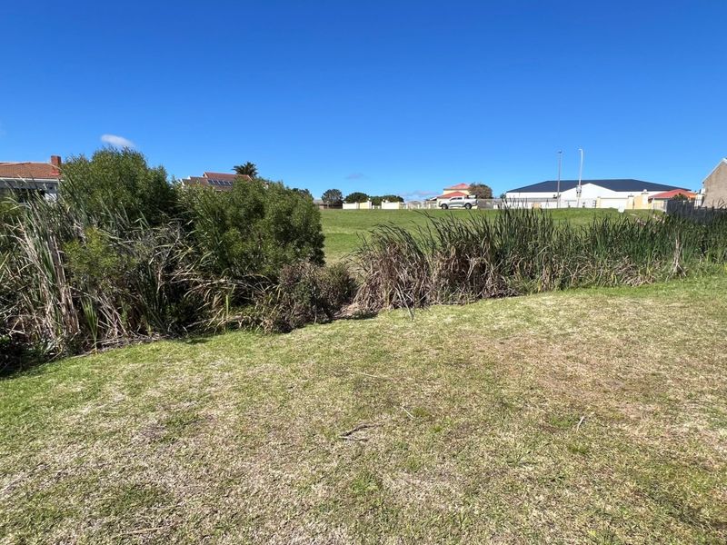 A view of a vacant residential plot featuring a grass lawn and natural vegetation, situated in a developed neighborhood.