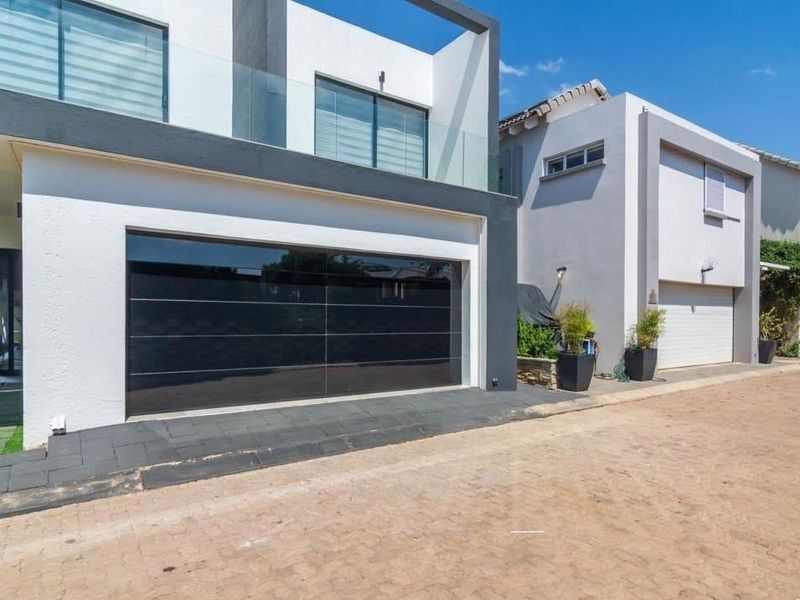 Modern double-story residential facade featuring a sleek black garage door, glass balcony railing, and paved driveway in a contemporary estate setting.