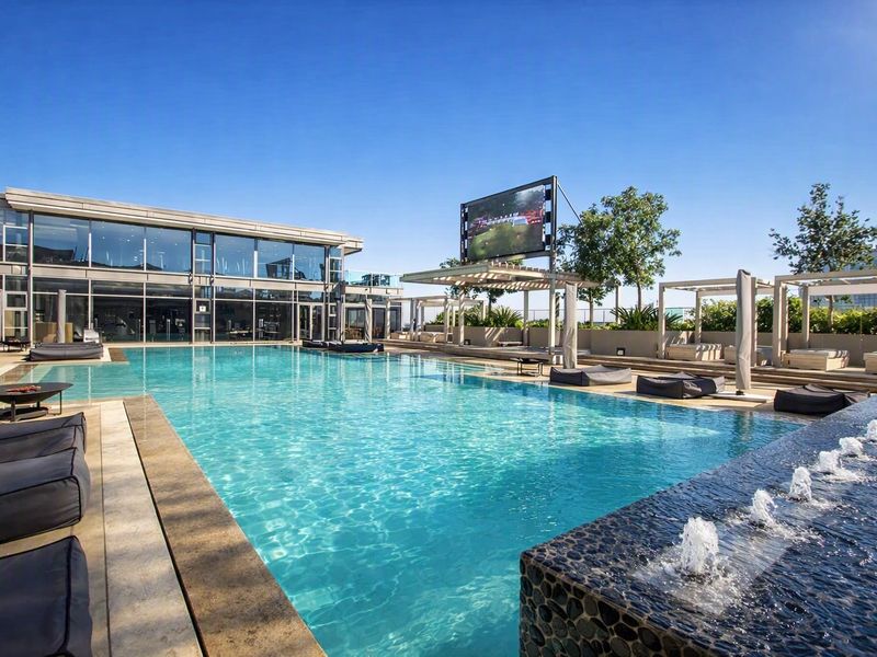 A modern, well-maintained swimming pool area featuring a water feature, pergolas, and a large glass-fronted building, perfect for outdoor leisure and recreation.