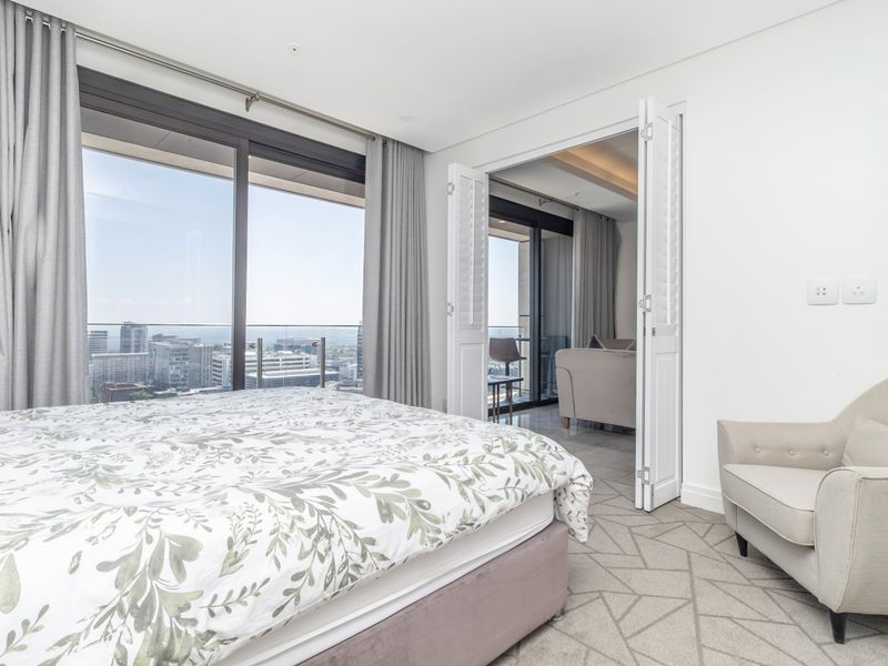 A bright, modern bedroom featuring large sliding glass doors leading to a balcony with city views and elegant double shutter doors.