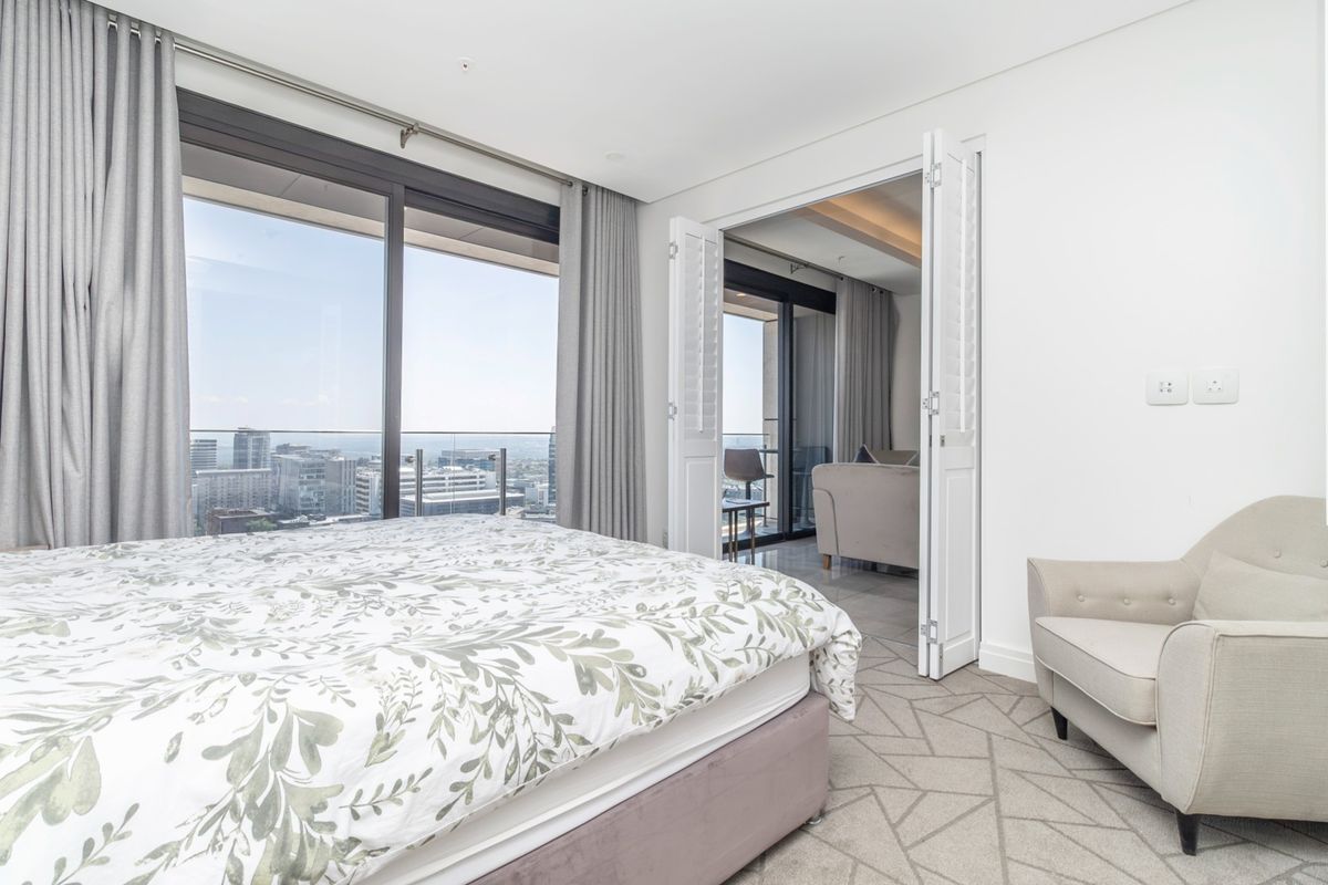 A bright, modern bedroom featuring large sliding glass doors leading to a balcony with city views and elegant double shutter doors.