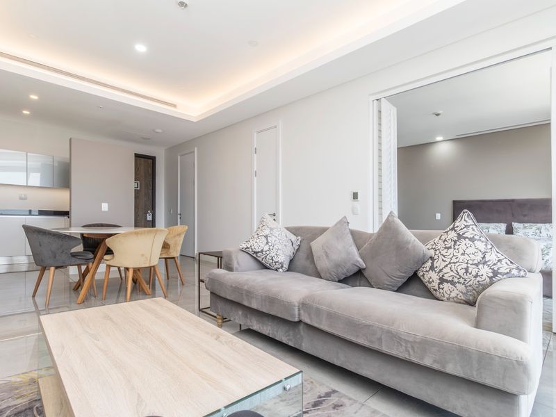 Modern open-plan living area featuring tiled floors, recessed ceiling lighting, and a sleek kitchen with stone countertops, seamlessly connected to an adjacent bedroom.