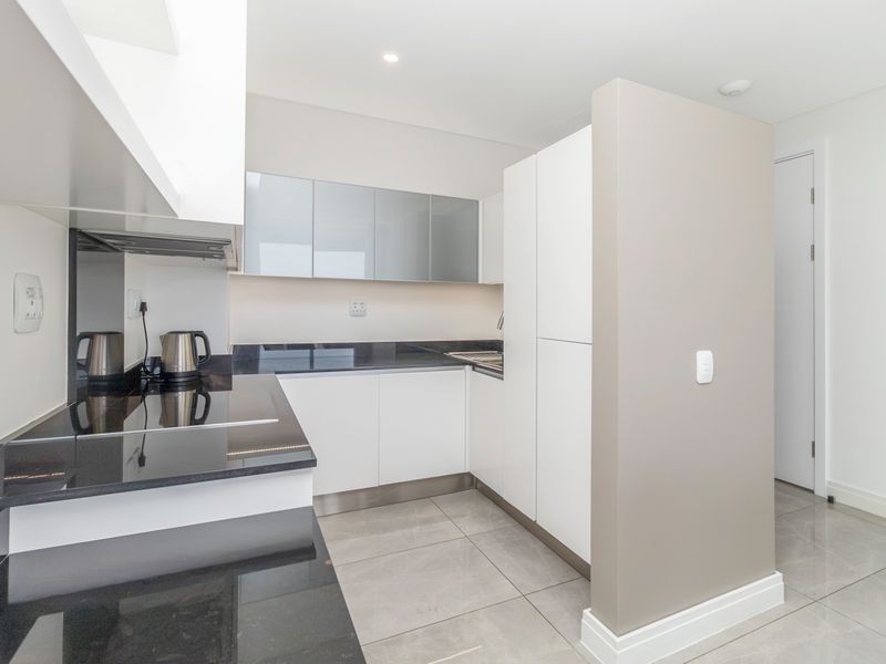 A modern, minimalist kitchen featuring sleek white cabinetry, dark granite countertops, integrated appliances, and neutral tiled flooring in excellent condition.