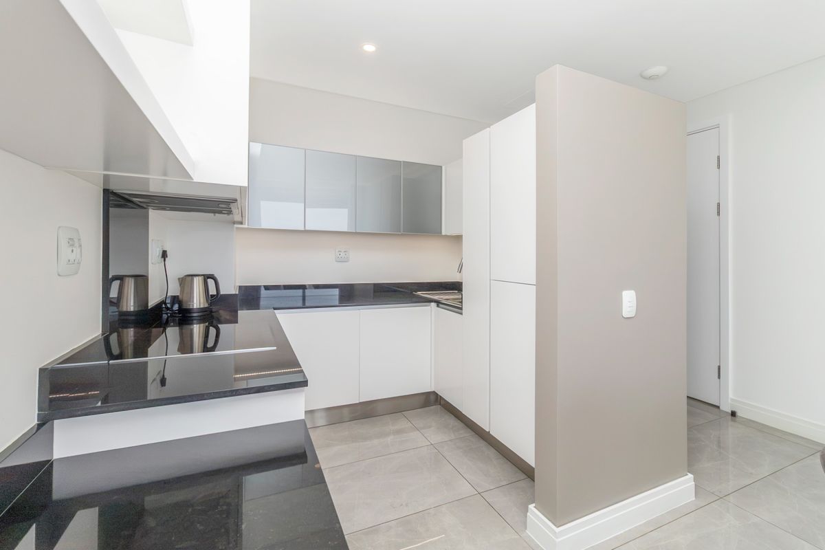 A modern, minimalist kitchen featuring sleek white cabinetry, dark granite countertops, integrated appliances, and neutral tiled flooring in excellent condition.