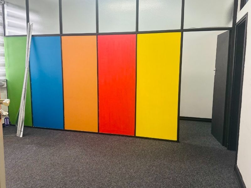 An empty office space featuring a multi-colored glass partition wall, grey carpet flooring, and standard office lighting.