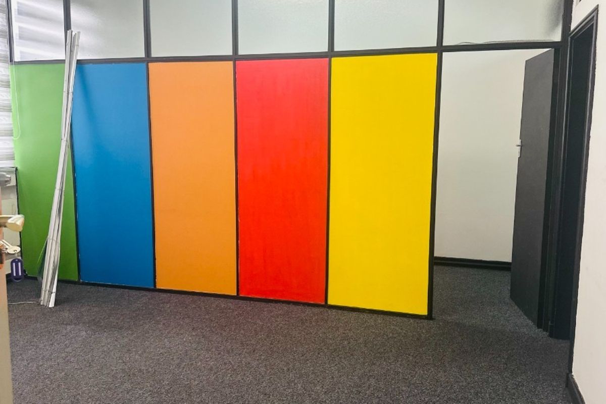 An empty office space featuring a multi-colored glass partition wall, grey carpet flooring, and standard office lighting.