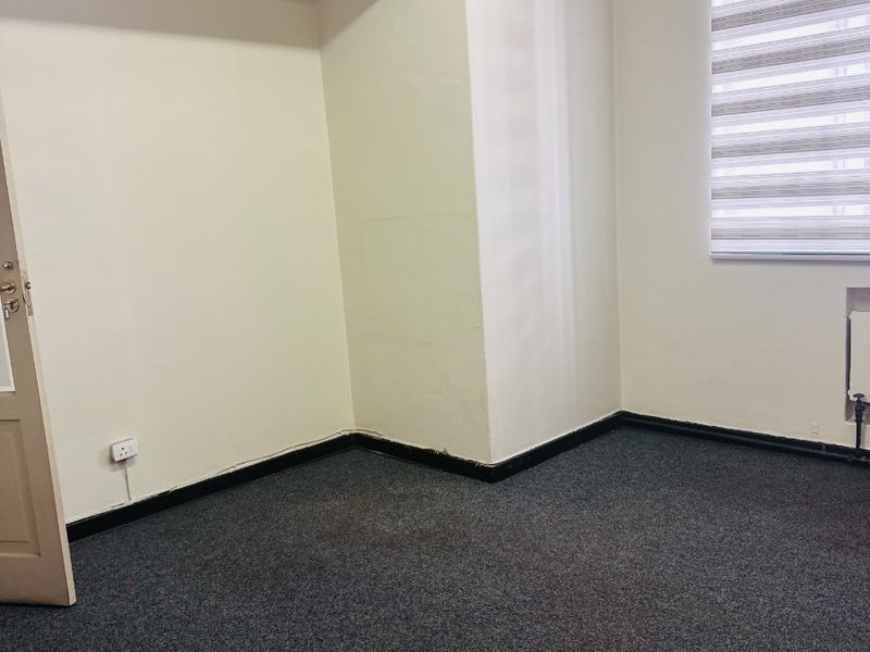 An empty office space featuring grey carpet flooring, neutral walls, and a window with a fitted roller blind.
