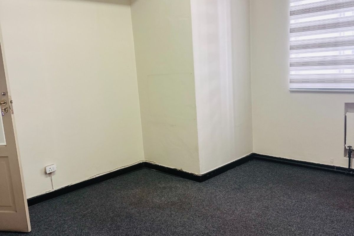 An empty office space featuring grey carpet flooring, neutral walls, and a window with a fitted roller blind.