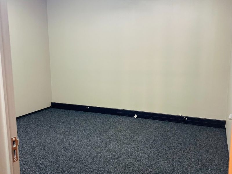 A clean, empty office space featuring grey carpet flooring, exposed ventilation ductwork, and functional overhead lighting.