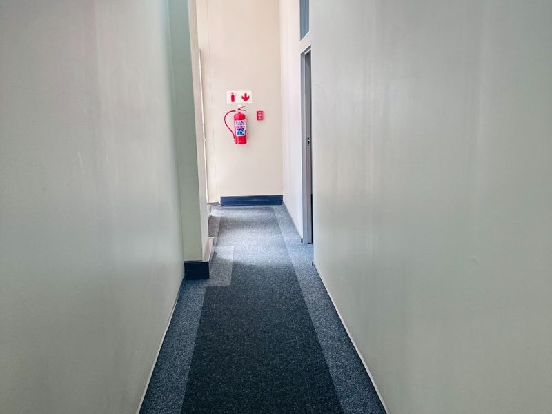 A functional office hallway featuring carpeted flooring, exposed HVAC ducting, and a wall-mounted fire extinguisher.