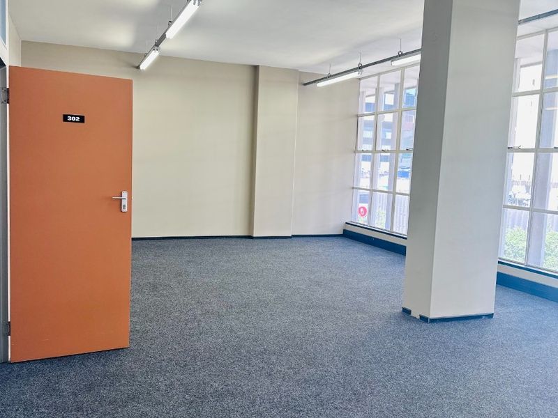A bright, open-plan office space featuring large windows, neutral walls, carpeted flooring, and functional overhead strip lighting.