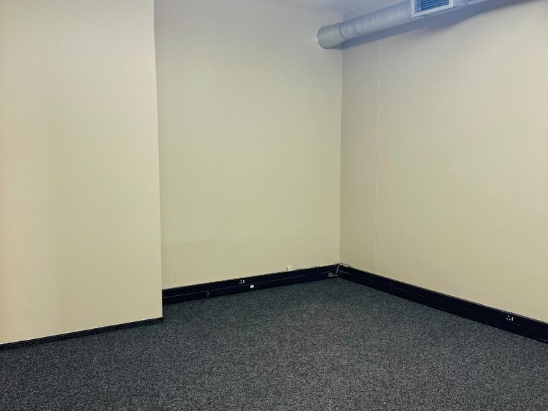 An empty office space featuring exposed industrial ductwork, fluorescent lighting, and grey carpeting, suitable for a professional workspace setup.