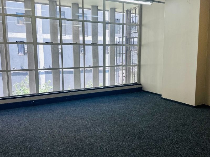 A bright, empty office space featuring large industrial-style windows, fitted carpeting, and overhead lighting tracks, suitable for a professional workspace.