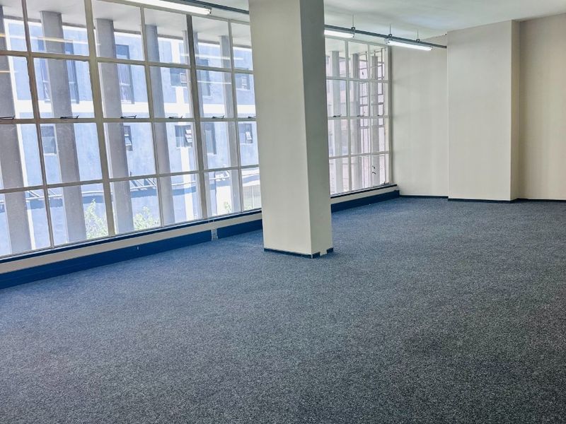A bright, open-plan office space featuring large windows, neutral carpeting, and functional overhead lighting, offering a versatile workspace ready for customization.