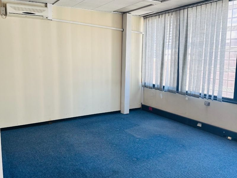 An empty office space featuring a suspended ceiling, wall-mounted air conditioning, large windows with vertical blinds, and blue carpet flooring.
