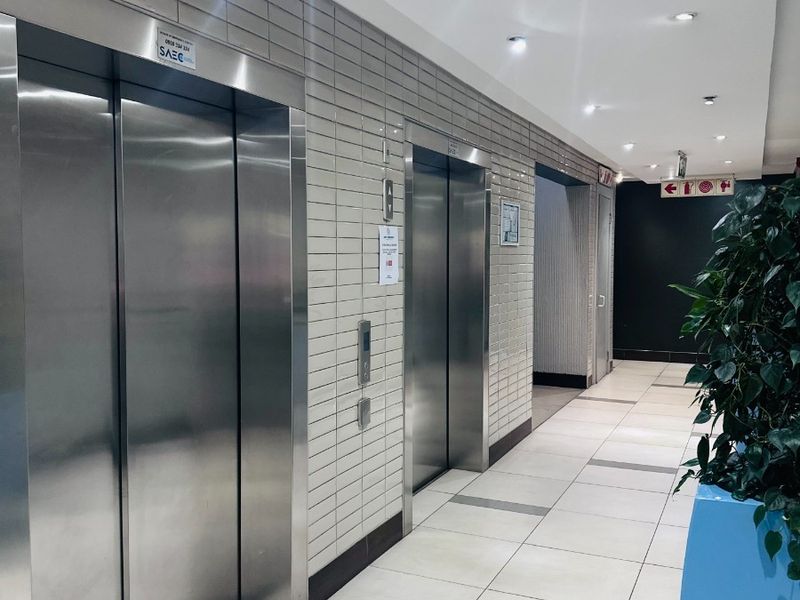 A clean, modern office building hallway featuring stainless steel elevator banks, tiled flooring, and recessed ceiling lighting.