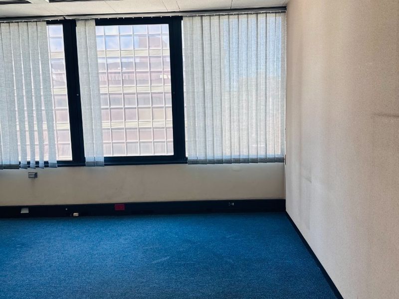 An empty office space featuring blue carpet flooring, a suspended ceiling, and large windows with vertical blinds, offering a blank canvas for professional use.