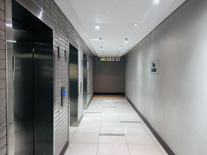 A clean and modern office building hallway featuring stainless steel elevator doors, tiled flooring, and recessed ceiling lighting.