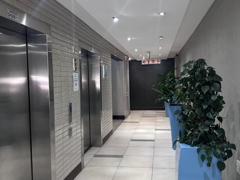 A modern office building hallway featuring stainless steel elevator doors, tiled flooring, and recessed ceiling lighting.