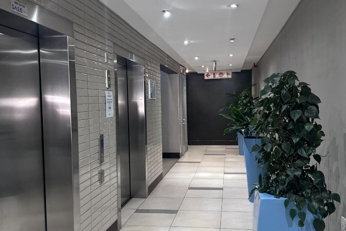 A modern office building hallway featuring stainless steel elevator doors, tiled flooring, and recessed ceiling lighting.