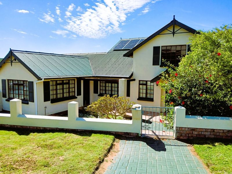 A charming front elevation of a house featuring a corrugated metal roof, solar panels, and a well-maintained garden with a paved entrance walkway.