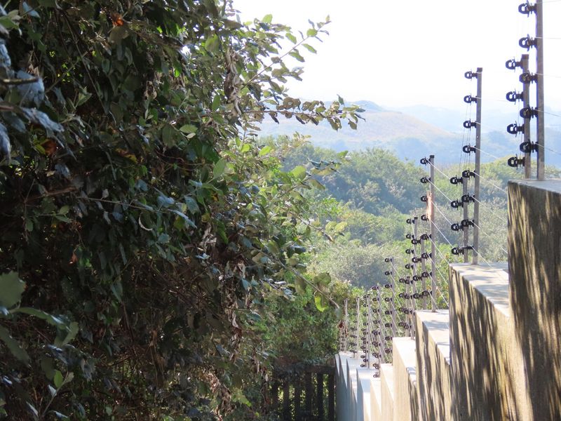 A view of the property boundary featuring a stepped retaining wall topped with electric security fencing, overlooking a lush green landscape.