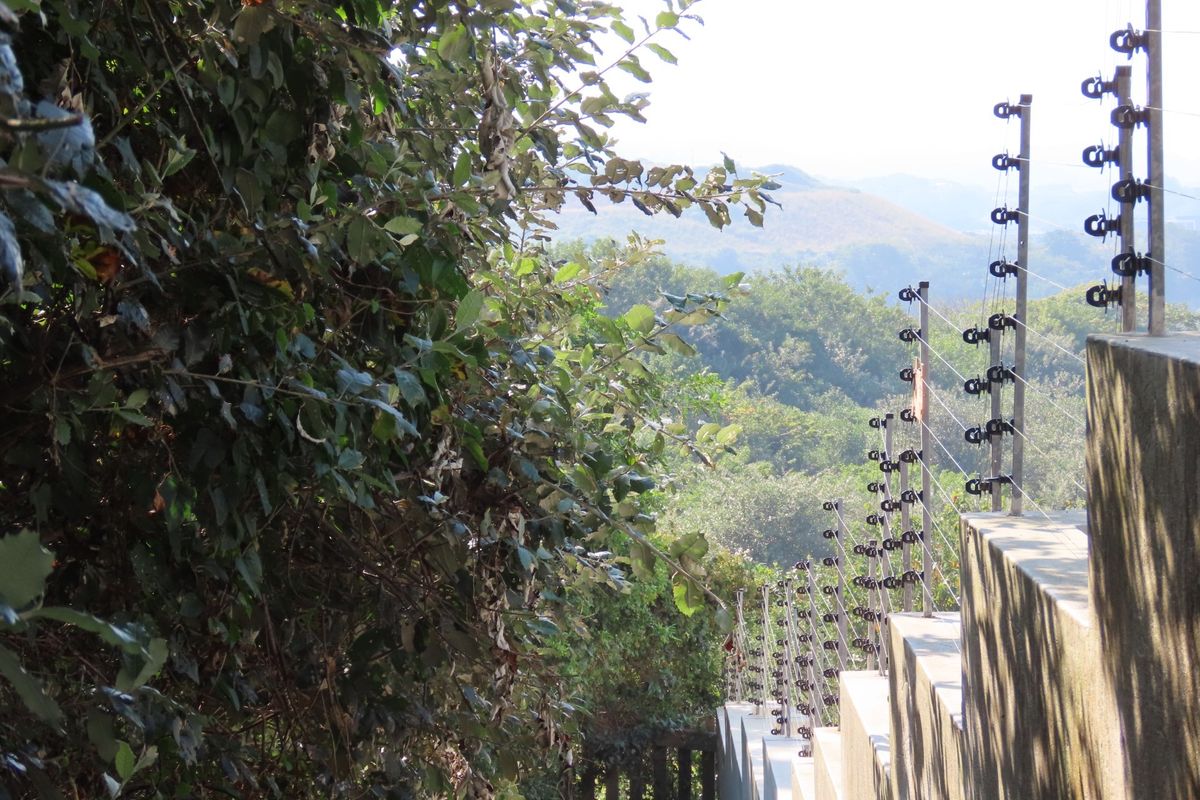A view of the property boundary featuring a stepped retaining wall topped with electric security fencing, overlooking a lush green landscape.