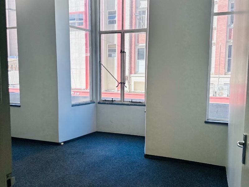 An empty office space featuring large windows, a suspended ceiling, and carpeted flooring, offering a bright and functional workspace.