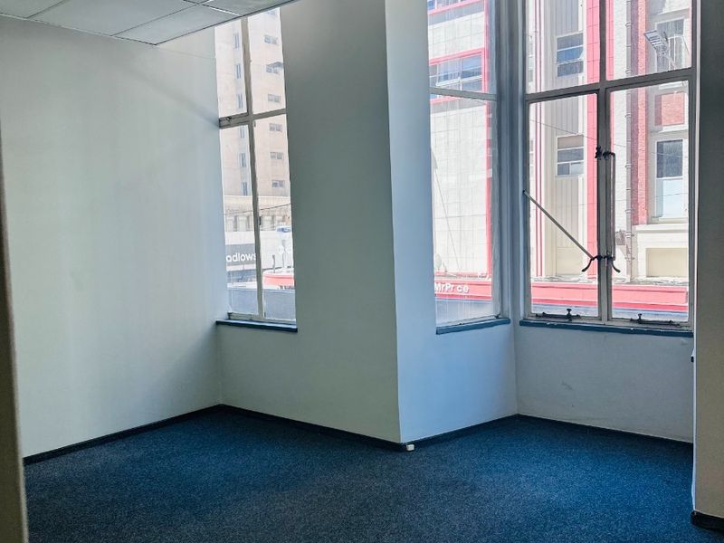 A vacant, well-lit office space featuring large windows, carpeted flooring, and a suspended ceiling with recessed lighting.