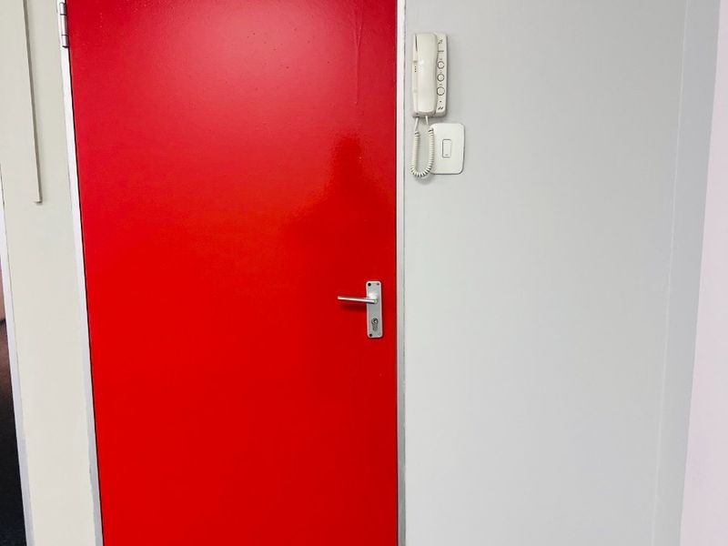 A functional office doorway featuring a red door with a magnetic lock, intercom system, and standard electrical fixtures.